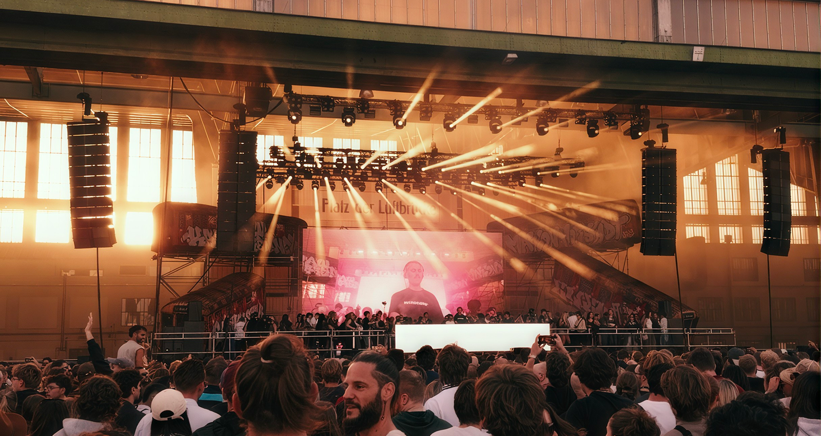 Crowd watching a live DJ performance on a stage with bright spotlights and large speakers inside an industrial-style venue with tall windows and a high ceiling.